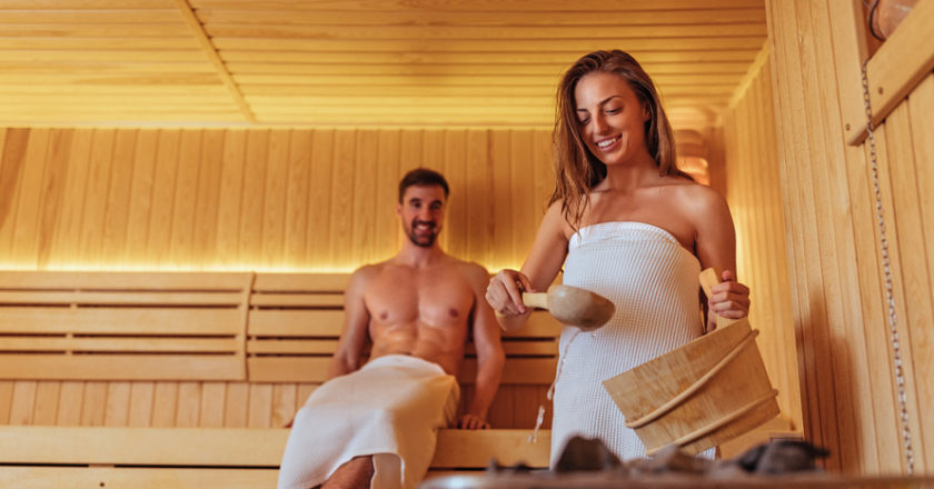 Frau macht Aufguss in der Sauna mit Mann im Hintergrund